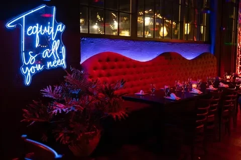 Dimly lit bar with red velvet booth, set tables, and a tequila sign.