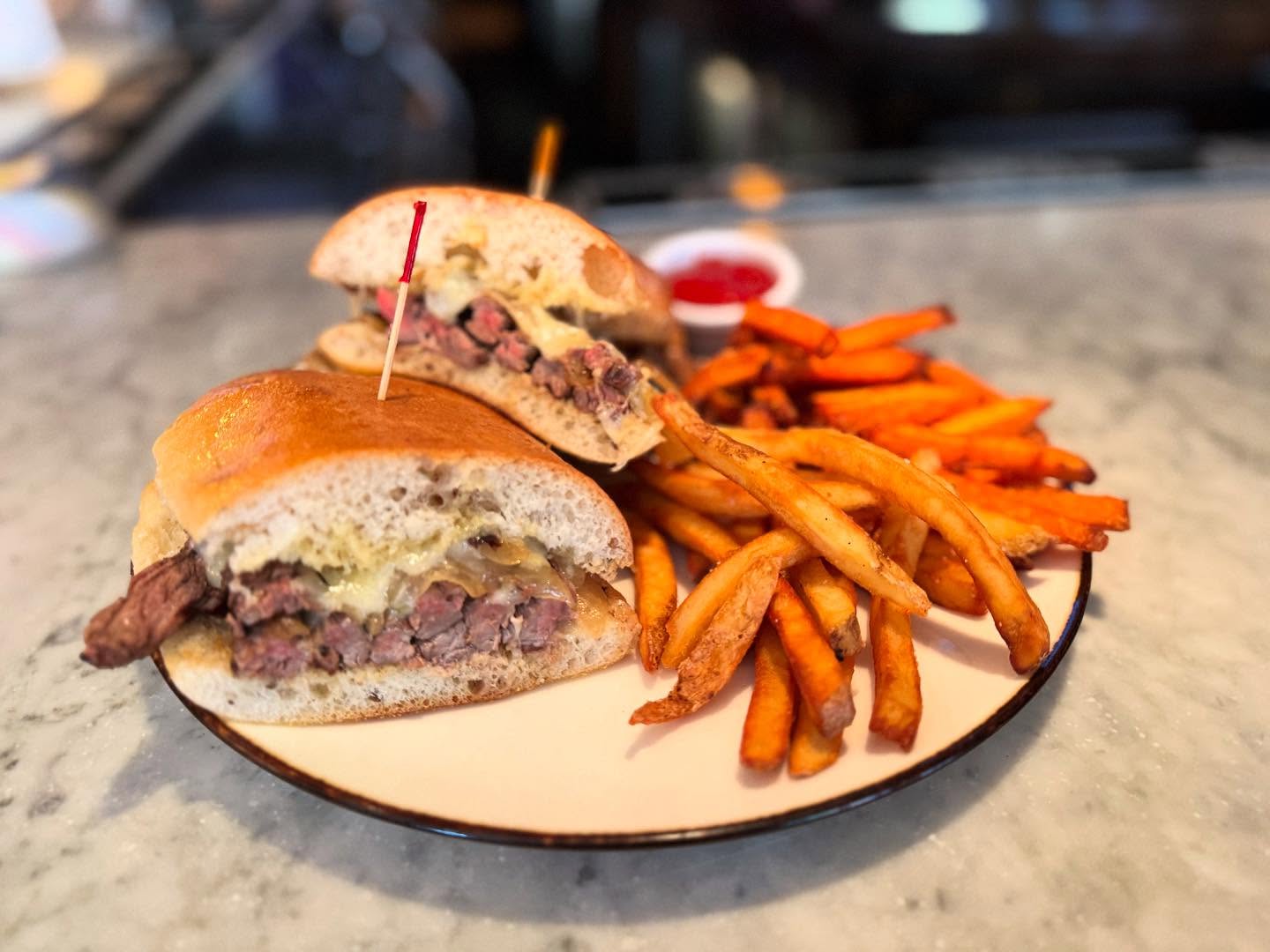 Steak and cheese sandwich with golden and orange fries on a plate.