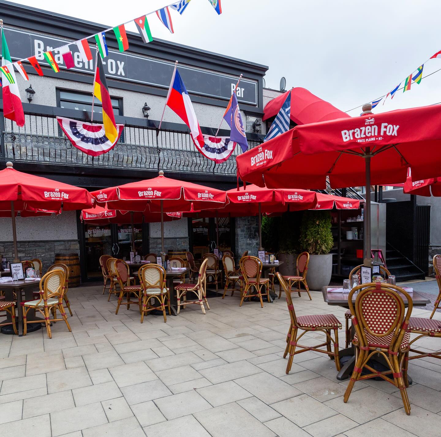 Outdoor dining at Brazen Fox with red umbrellas and international flags.