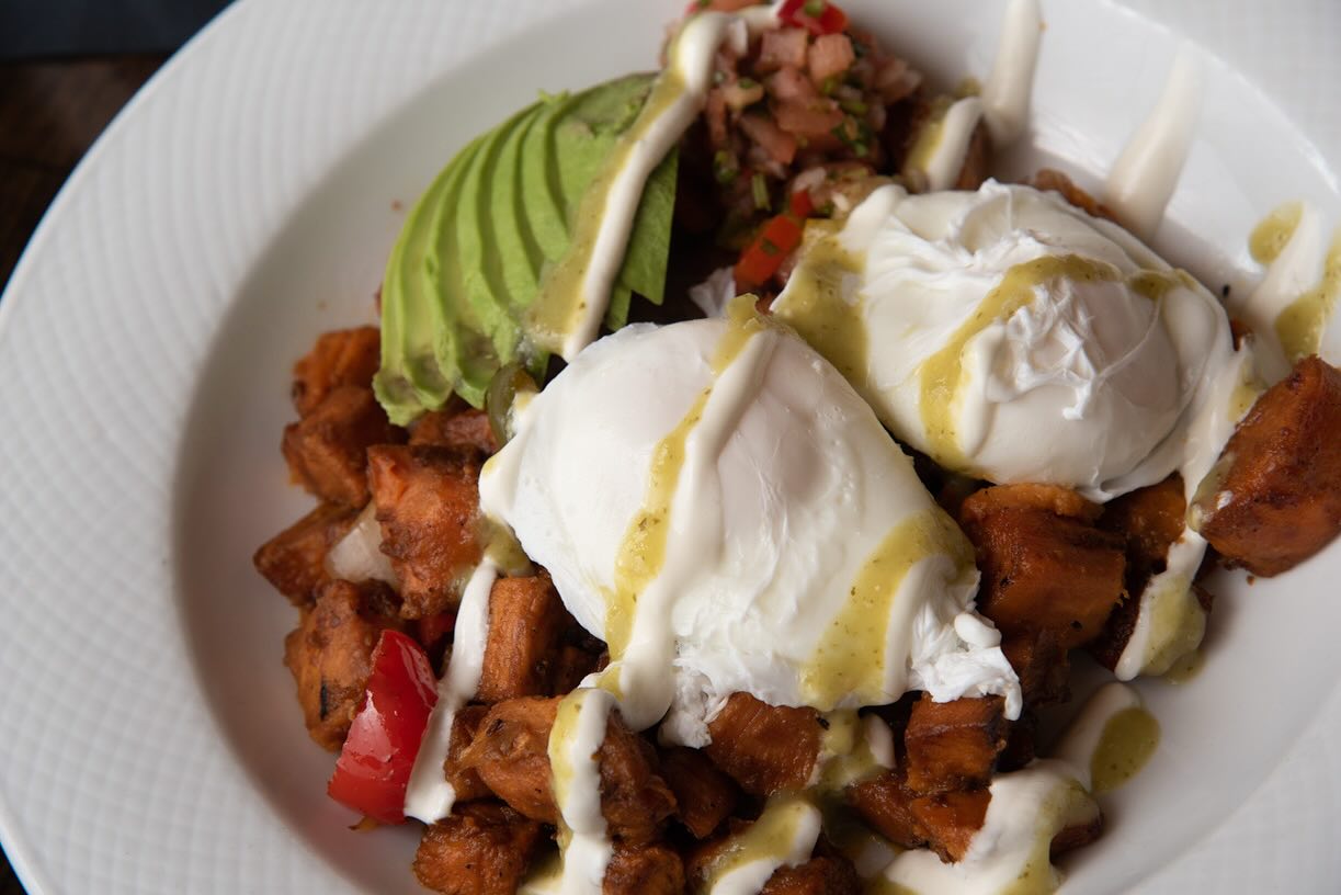 Poached eggs, avocado, sweet potato hash, and sauces in a white bowl.