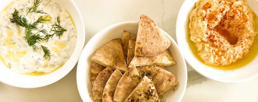 Tzatziki, pita bread, and hummus in white bowls on a white surface.