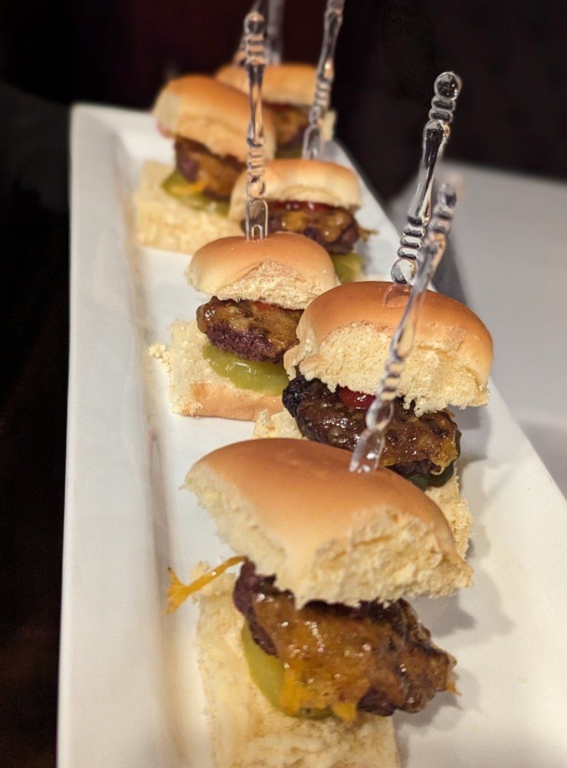 A row of cheeseburger sliders with cheese, pickles, and skewers.