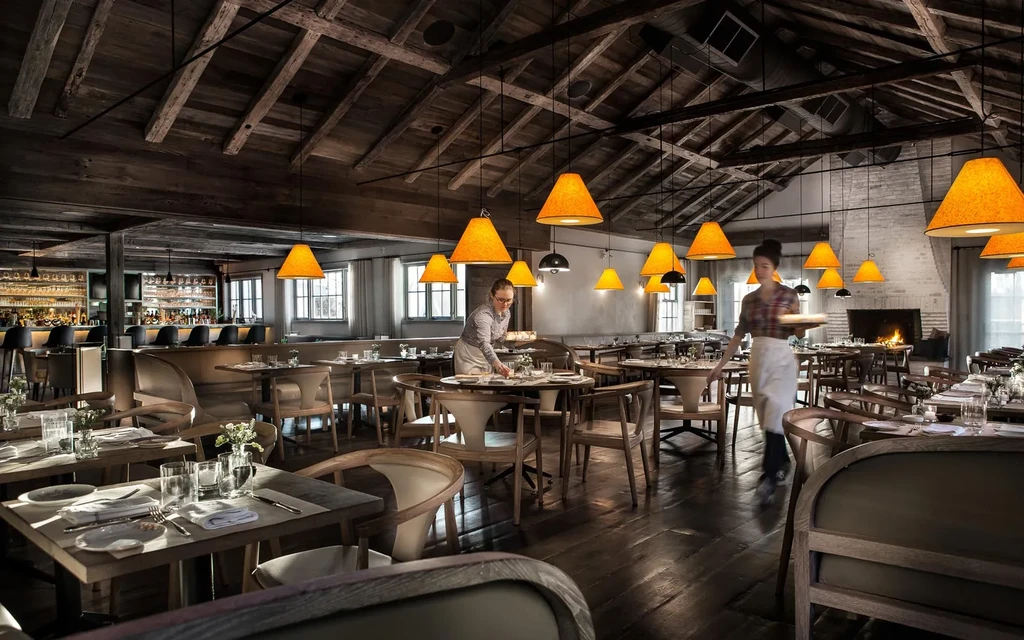 Two waitstaff prepare a rustic restaurant with orange lamps and wooden decor.