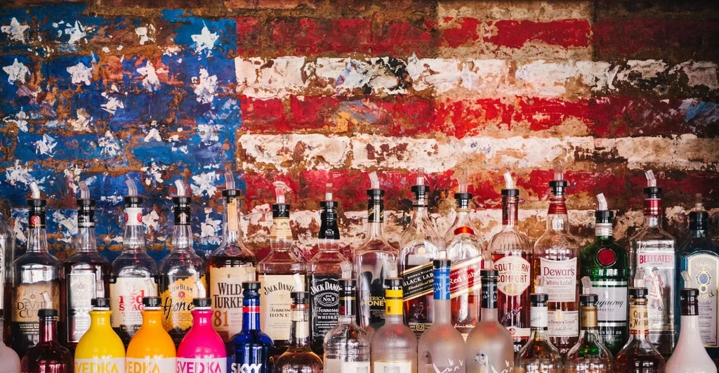 Assorted liquor bottles in front of a painted American flag on a brick wall.