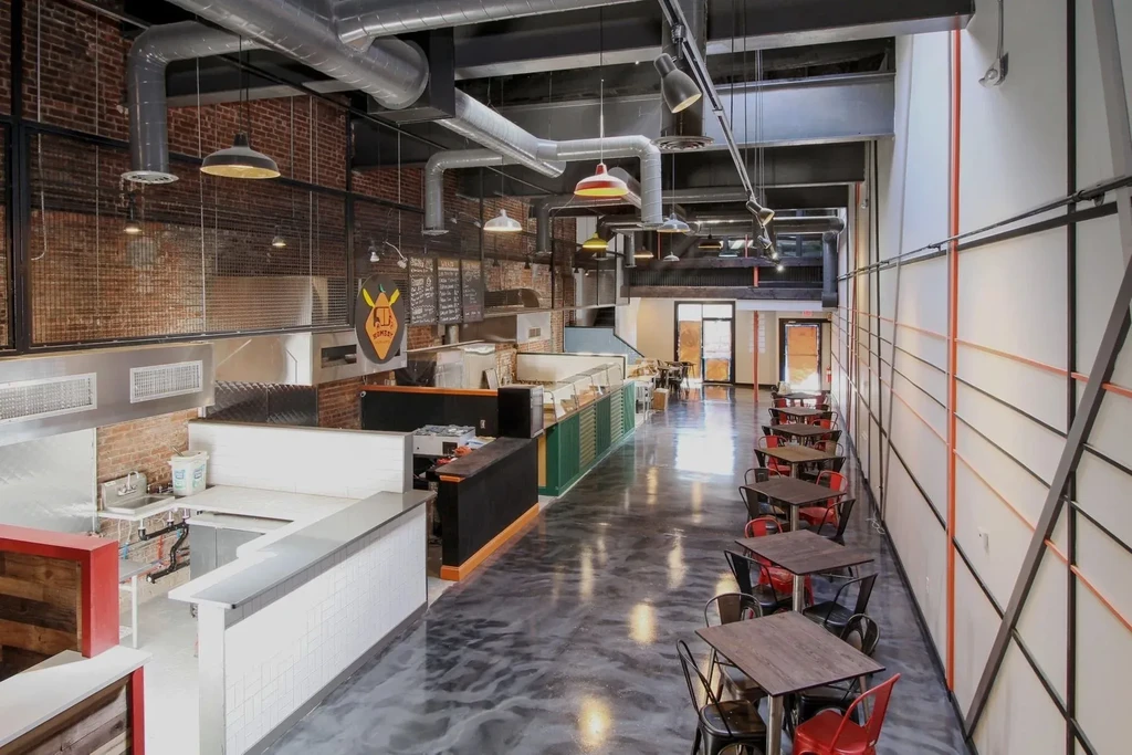High-ceiling industrial restaurant interior with exposed brick, ductwork, and open kitchen.