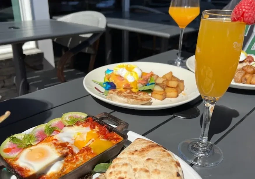 Breakfast with shakshuka, eggs Benedict, potatoes, and mimosas on an outdoor table.