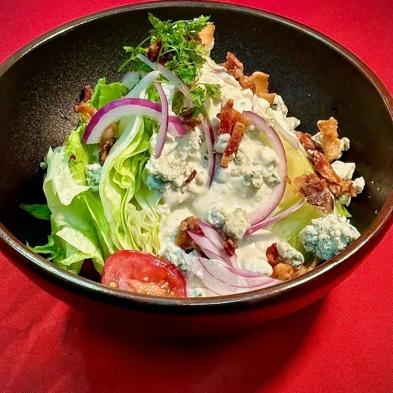 Wedge salad with blue cheese, bacon, red onion, and tomato in a black bowl.