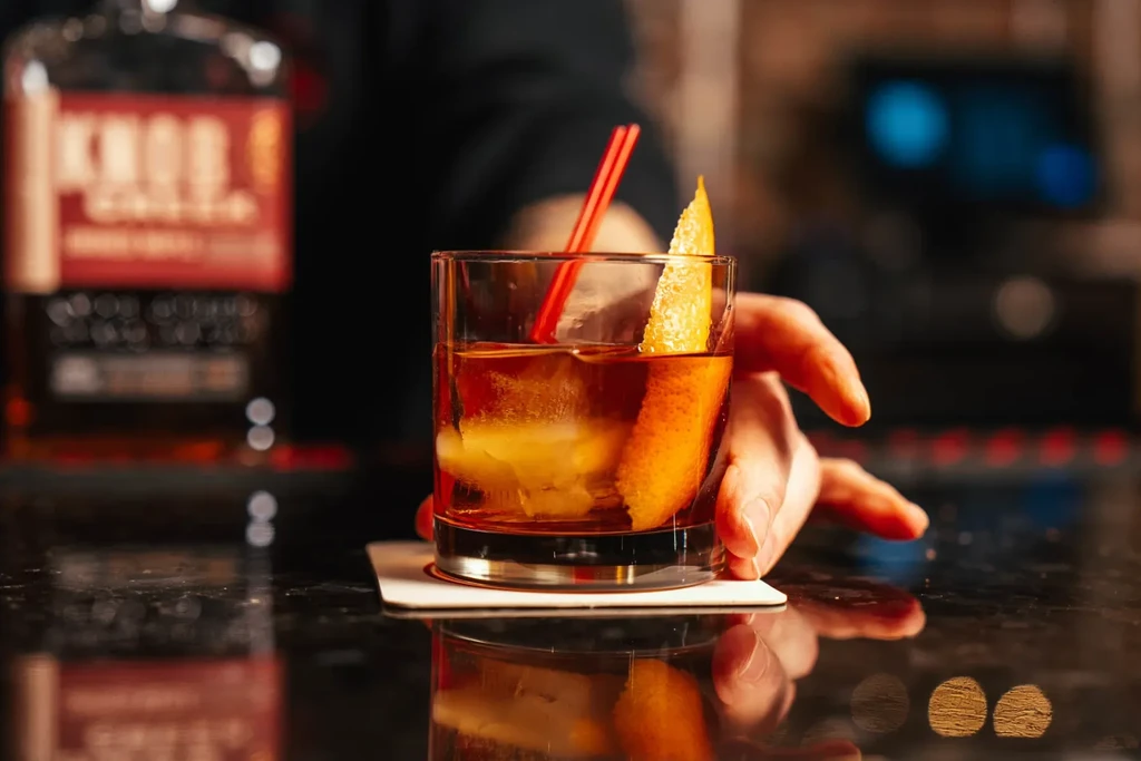 A hand reaches for a cocktail with ice, orange peel, and red straws.