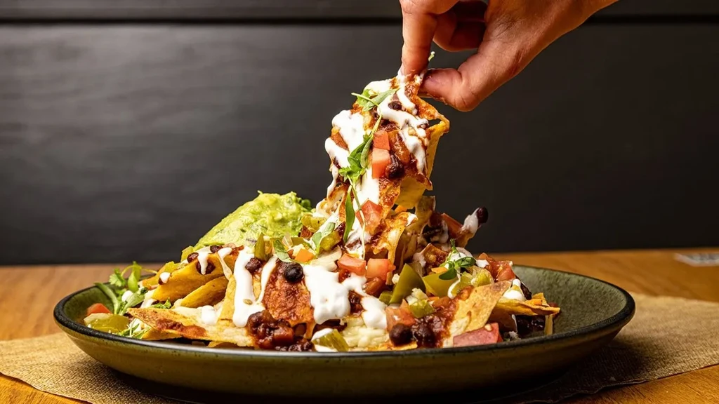 A hand lifts cheesy nachos from a plate with guacamole.