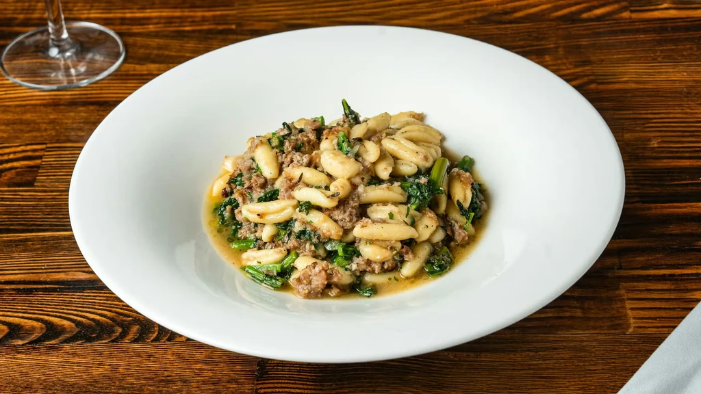 Overhead of ridged pasta, crumbled meat, and greens in white bowl.