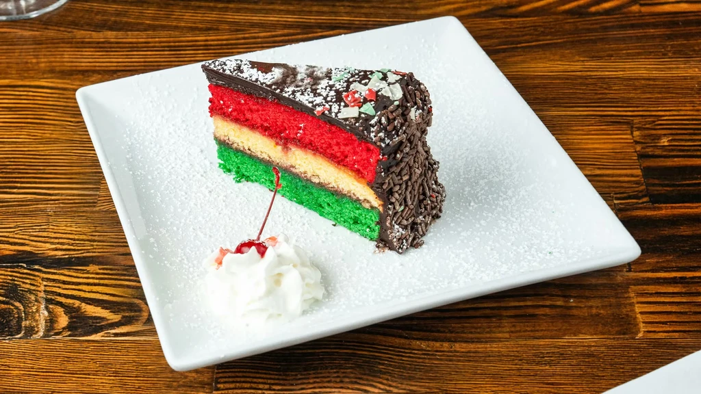Italian rainbow cookie cake slice with chocolate frosting, whipped cream, cherry.