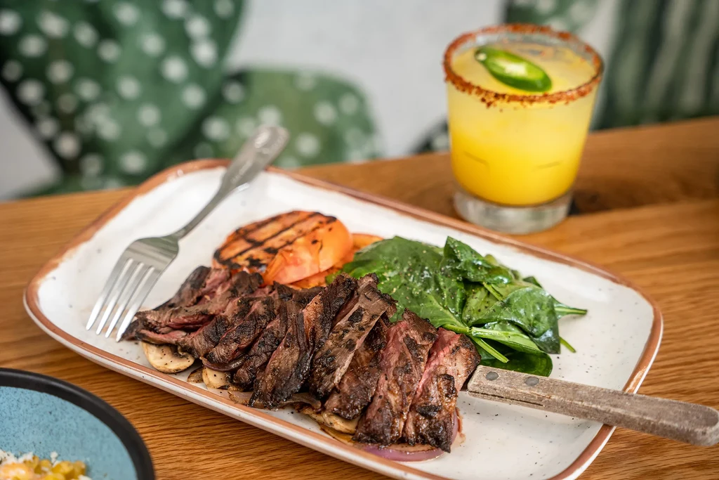 Sliced grilled steak, spinach, grilled tomato, and a margarita with a jalapeño slice.