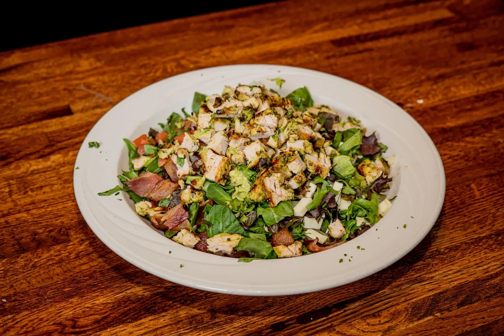 A white plate of chicken salad with bacon and greens on a wooden table.