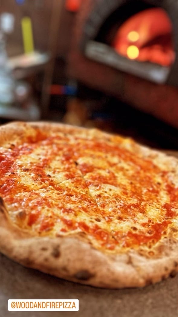 Cheese pizza in foreground, with a glowing wood-fired oven in background.