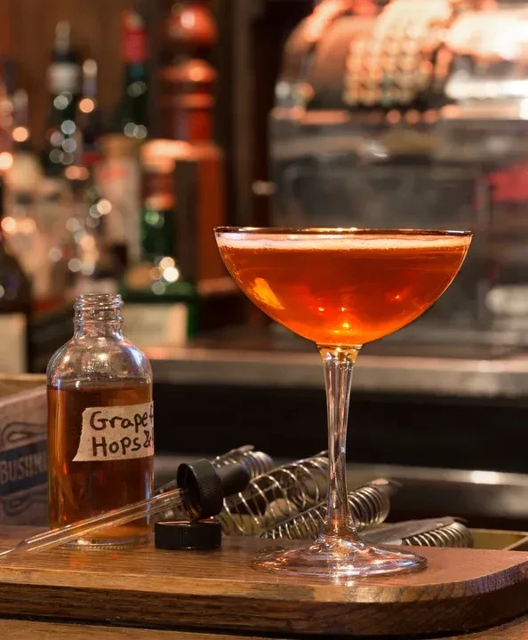 A coupe glass of amber cocktail and a dropper bottle of "Grape Hops".
