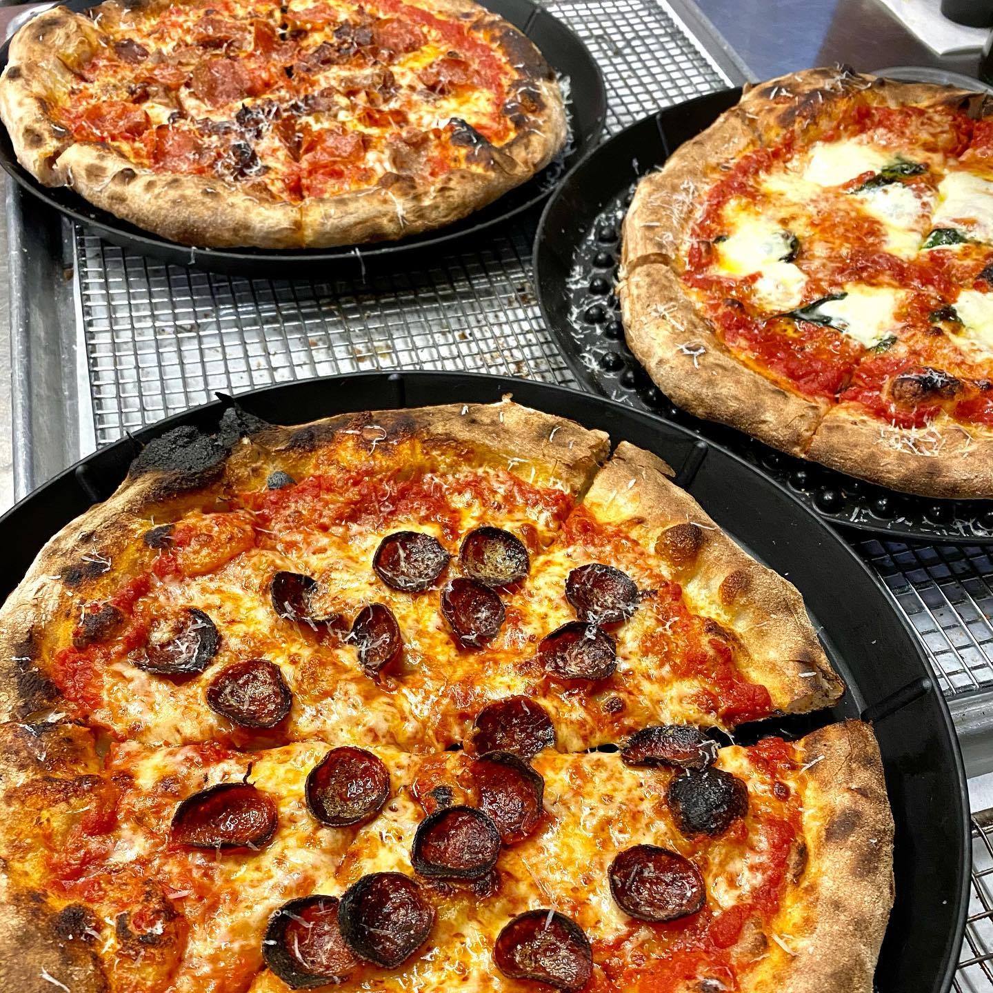 Three round pizzas, including pepperoni and margherita, on black pans.