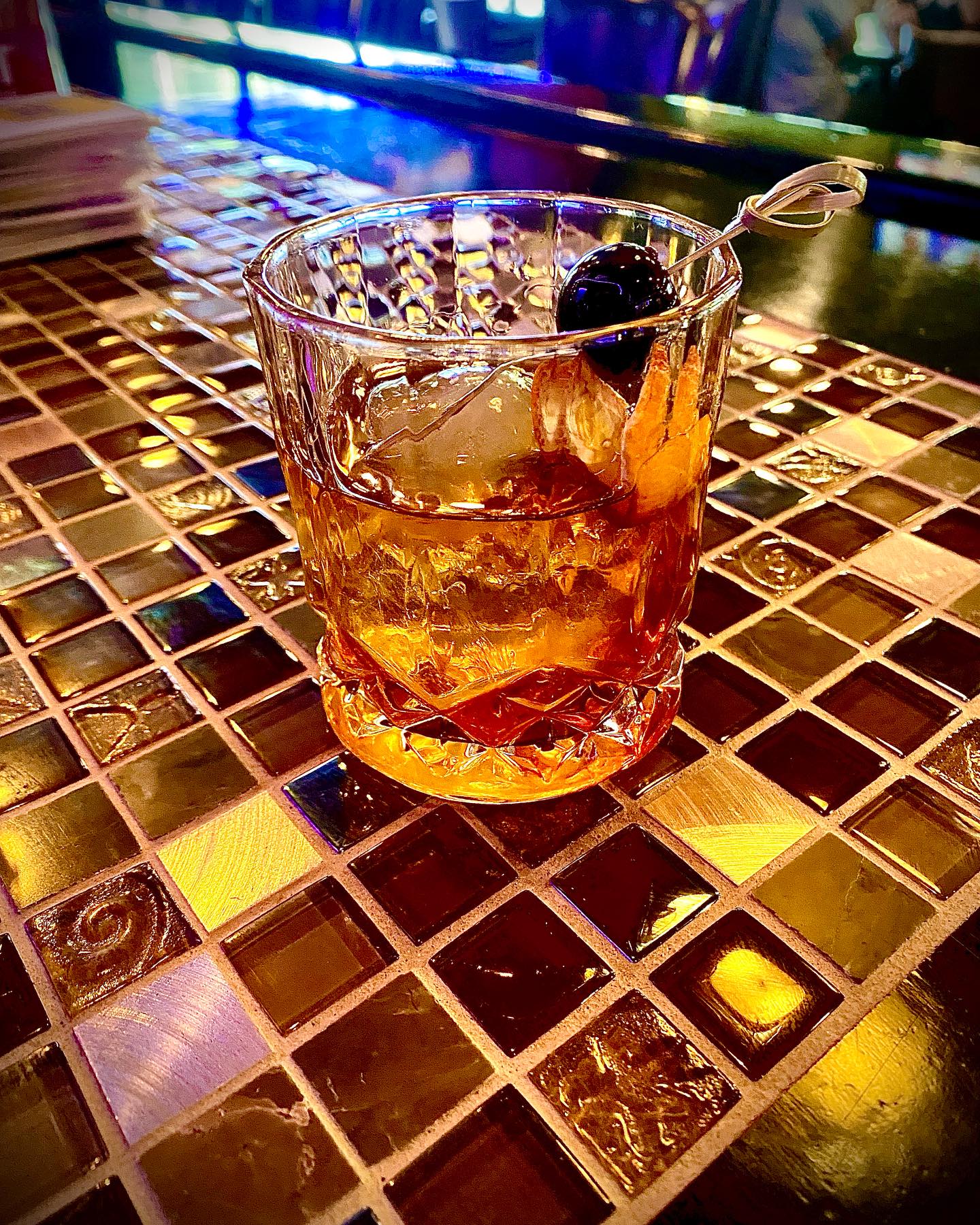 An amber cocktail with a cherry and ice on a mosaic tiled bar.