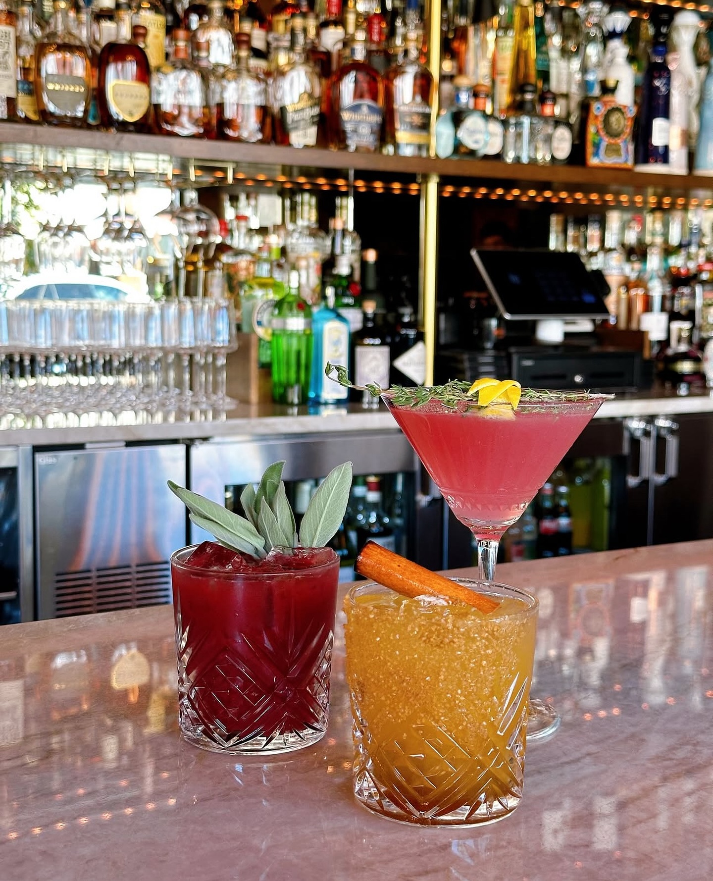 Three garnished cocktails on a bar counter, with liquor bottles behind.