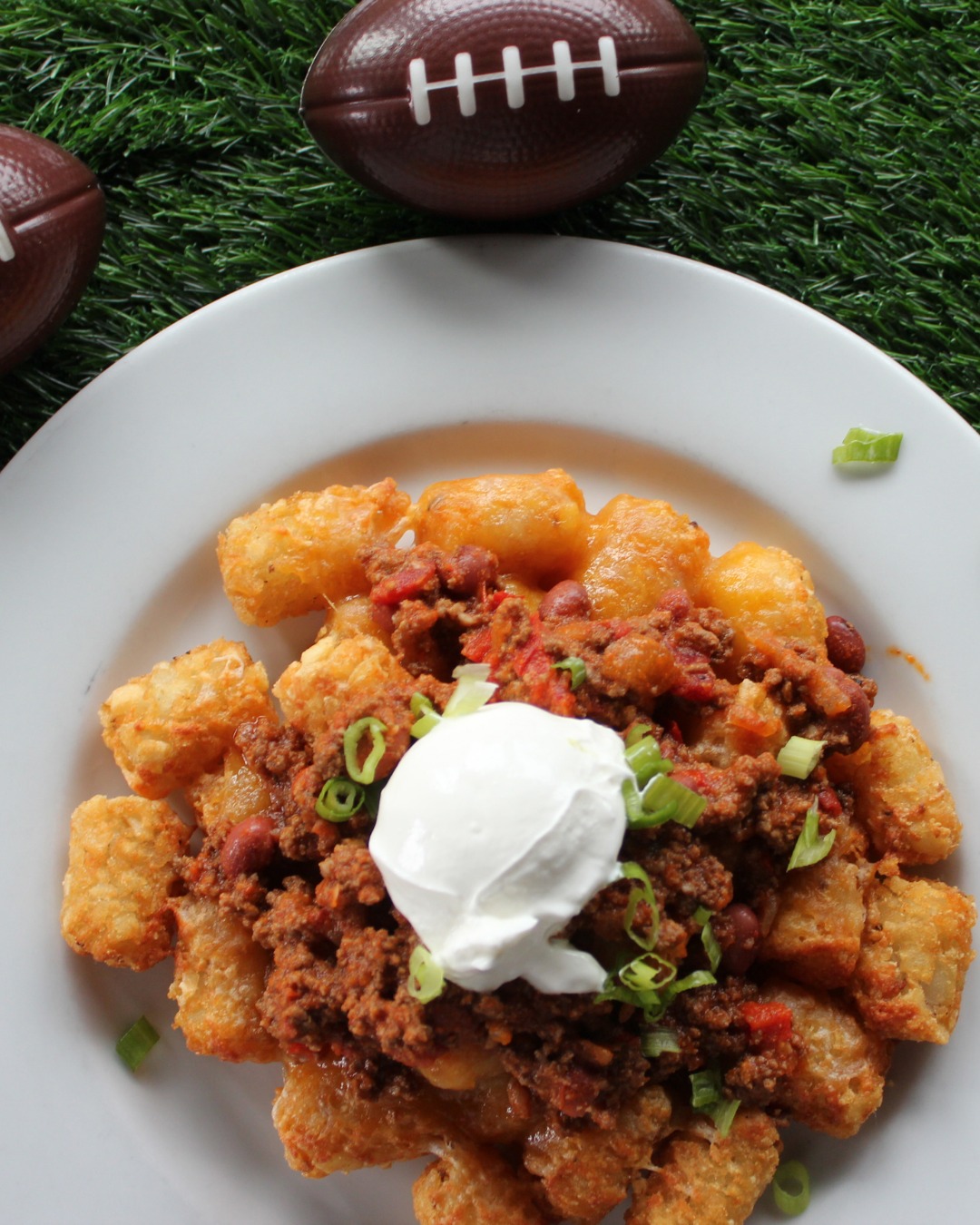 Chili tater tots with sour cream, green onions, and footballs on turf.