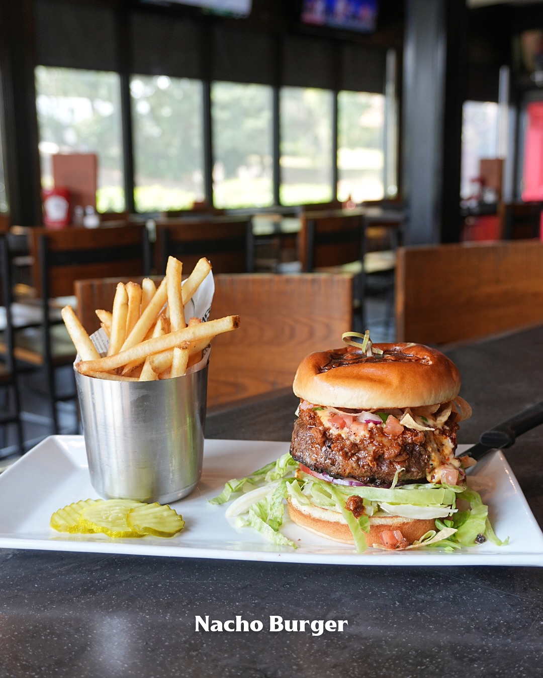 Nacho burger with fries and pickles on a white plate.