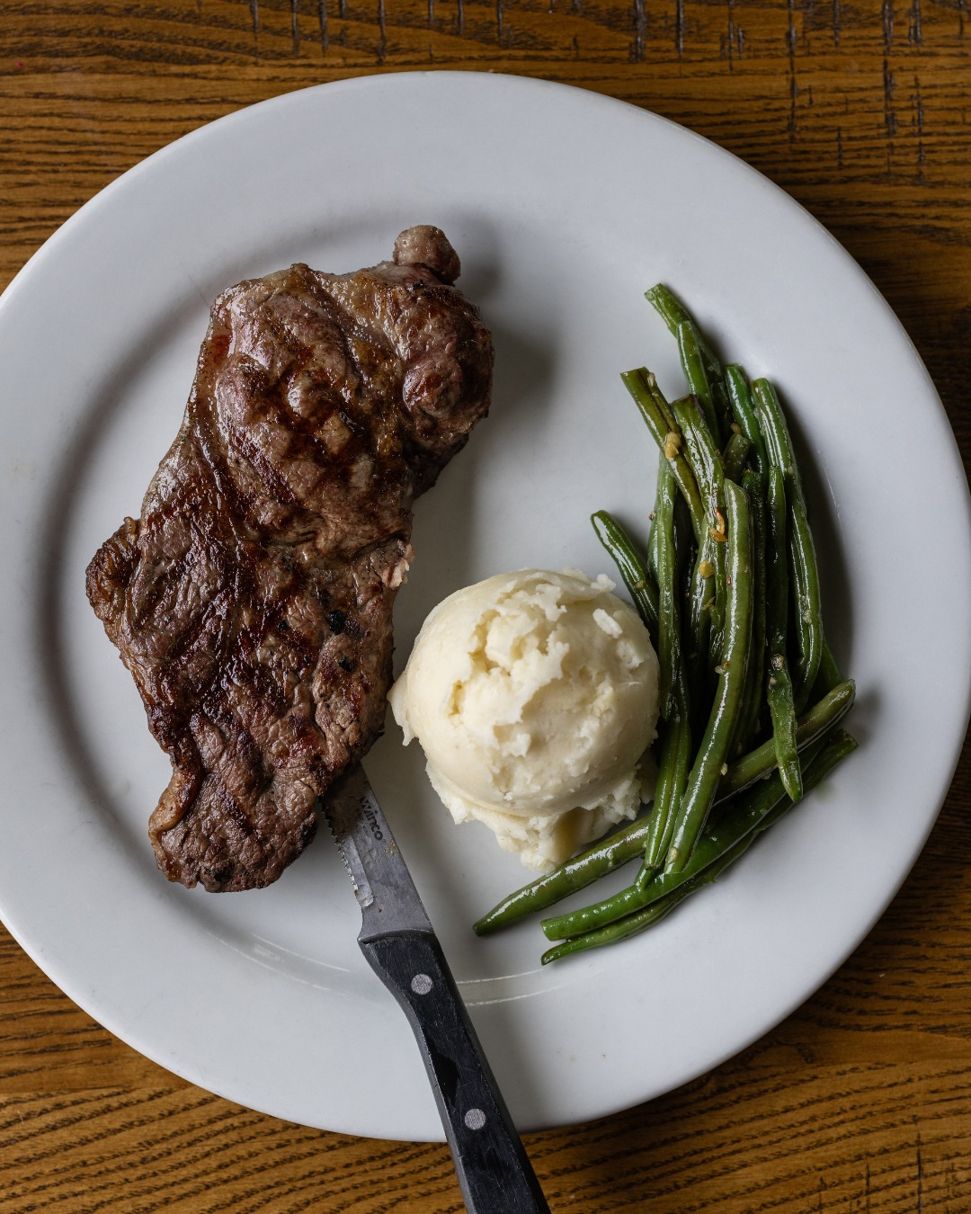 Steak, mashed potatoes, green beans, and a knife on a white plate.