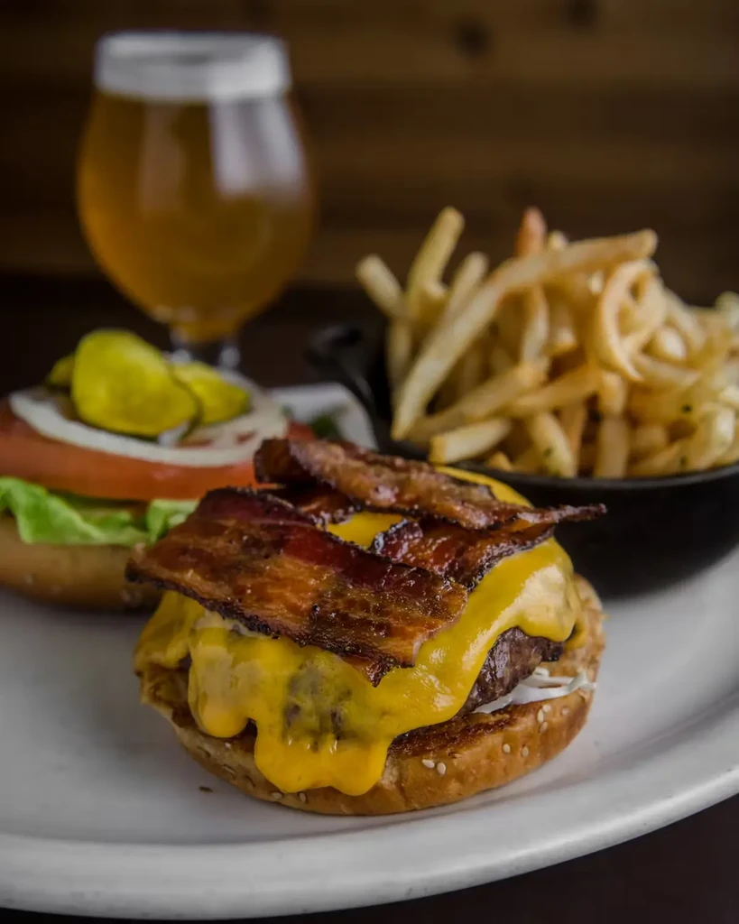 Cheeseburger with bacon, fries, and a beer on a white plate.