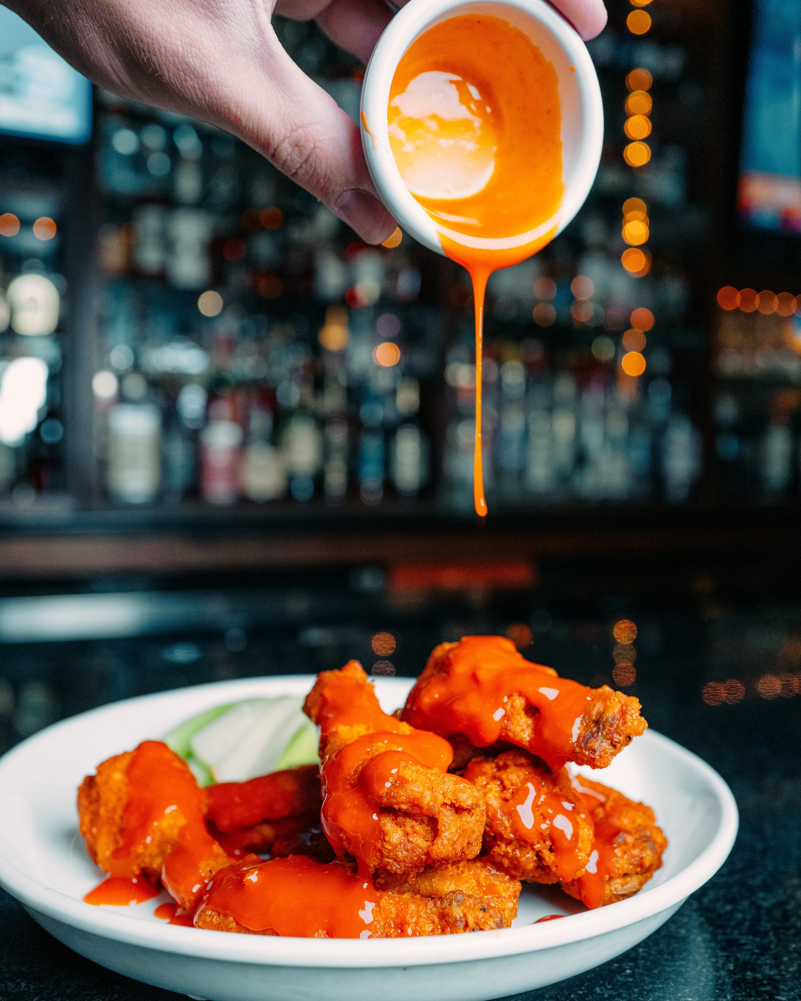 Hand pouring orange sauce over chicken wings at a bar.