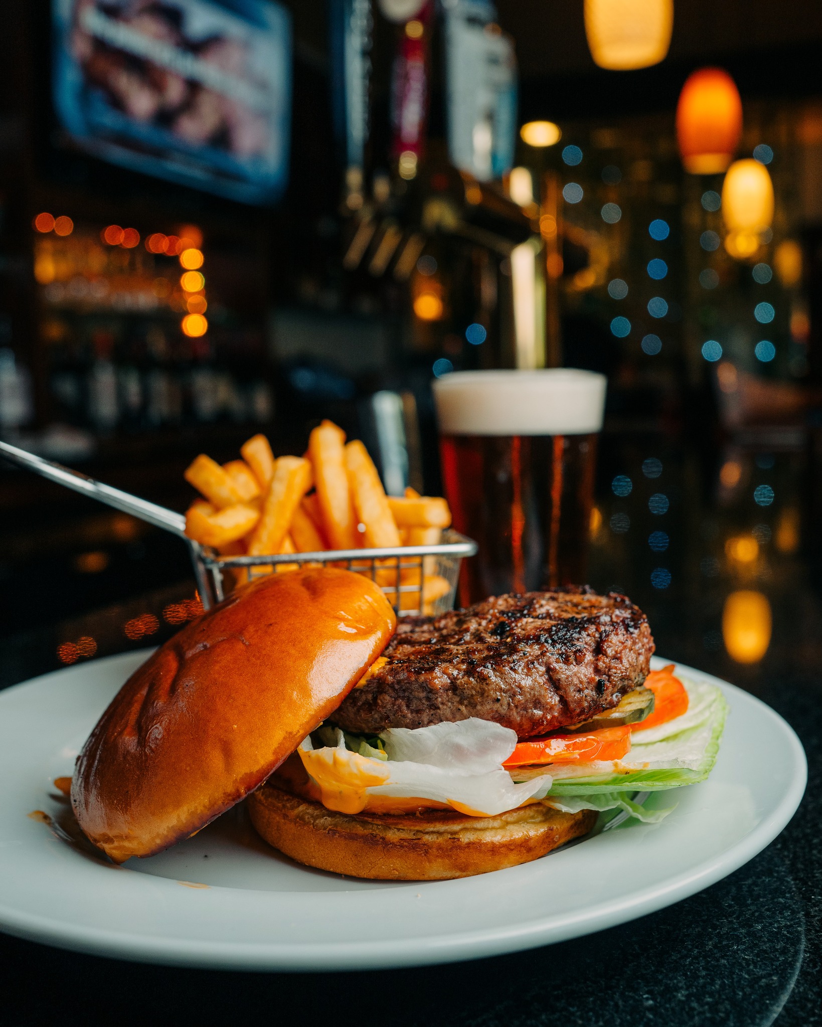 Plate with a burger, fries, and beer at a dimly lit bar.