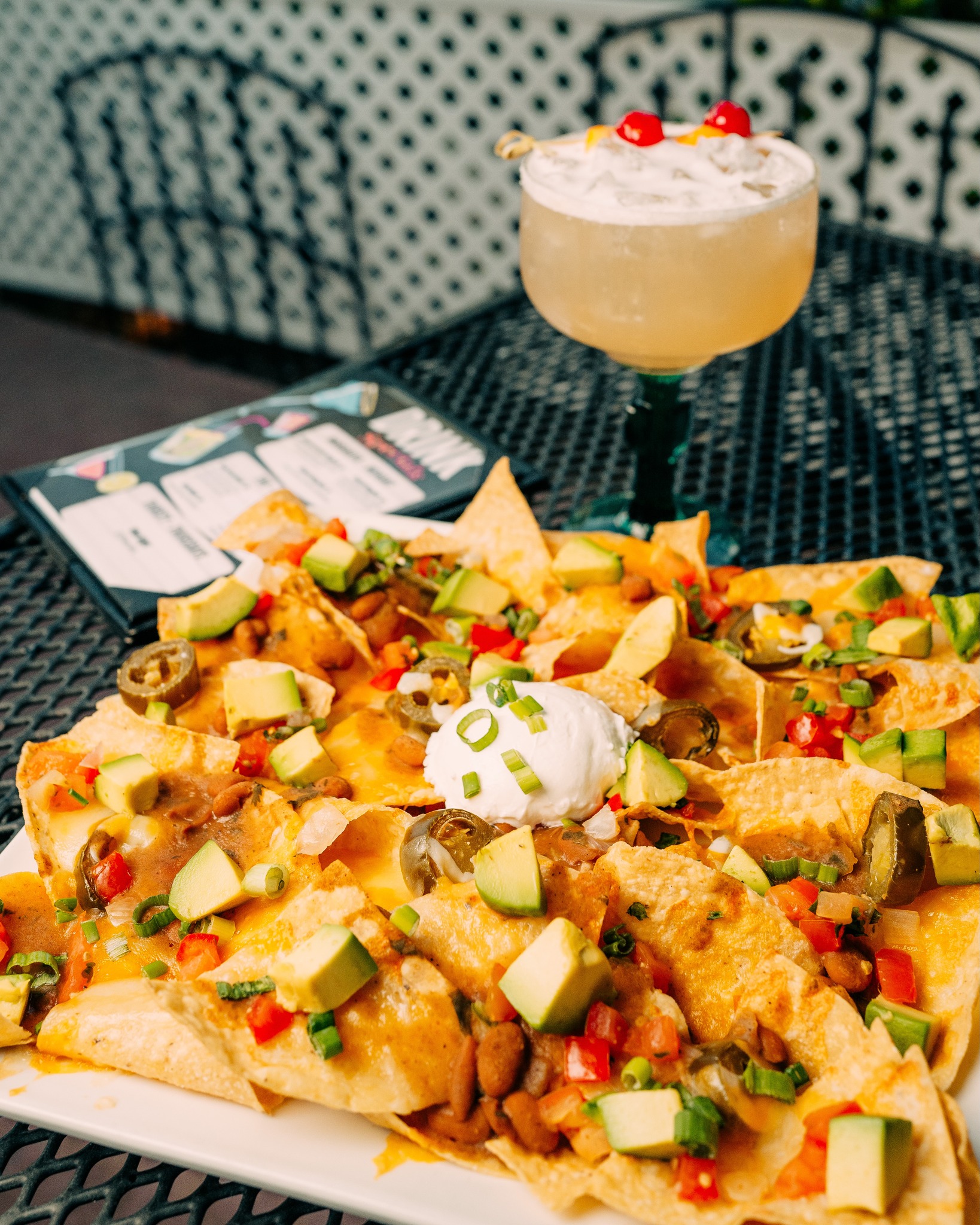 Nachos with avocado, jalapeños, sour cream, and a cocktail on a table.