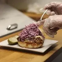 Gloved hands add purple coleslaw to a pulled pork sandwich on a white plate.