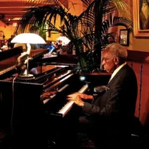 An elderly man in a suit plays a piano in a dimly lit room.