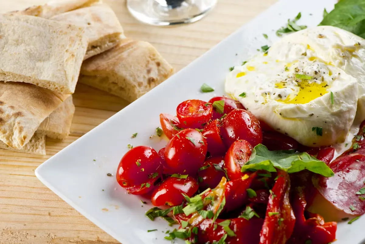 Burrata with sliced tomatoes, pita bread, and salami on a white plate.