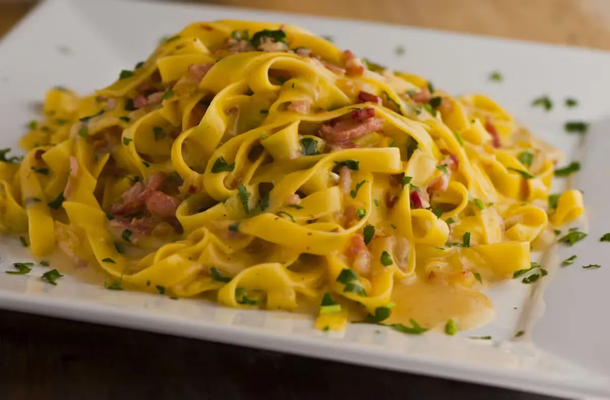 Yellow tagliatelle pasta with bacon and parsley on a white plate.