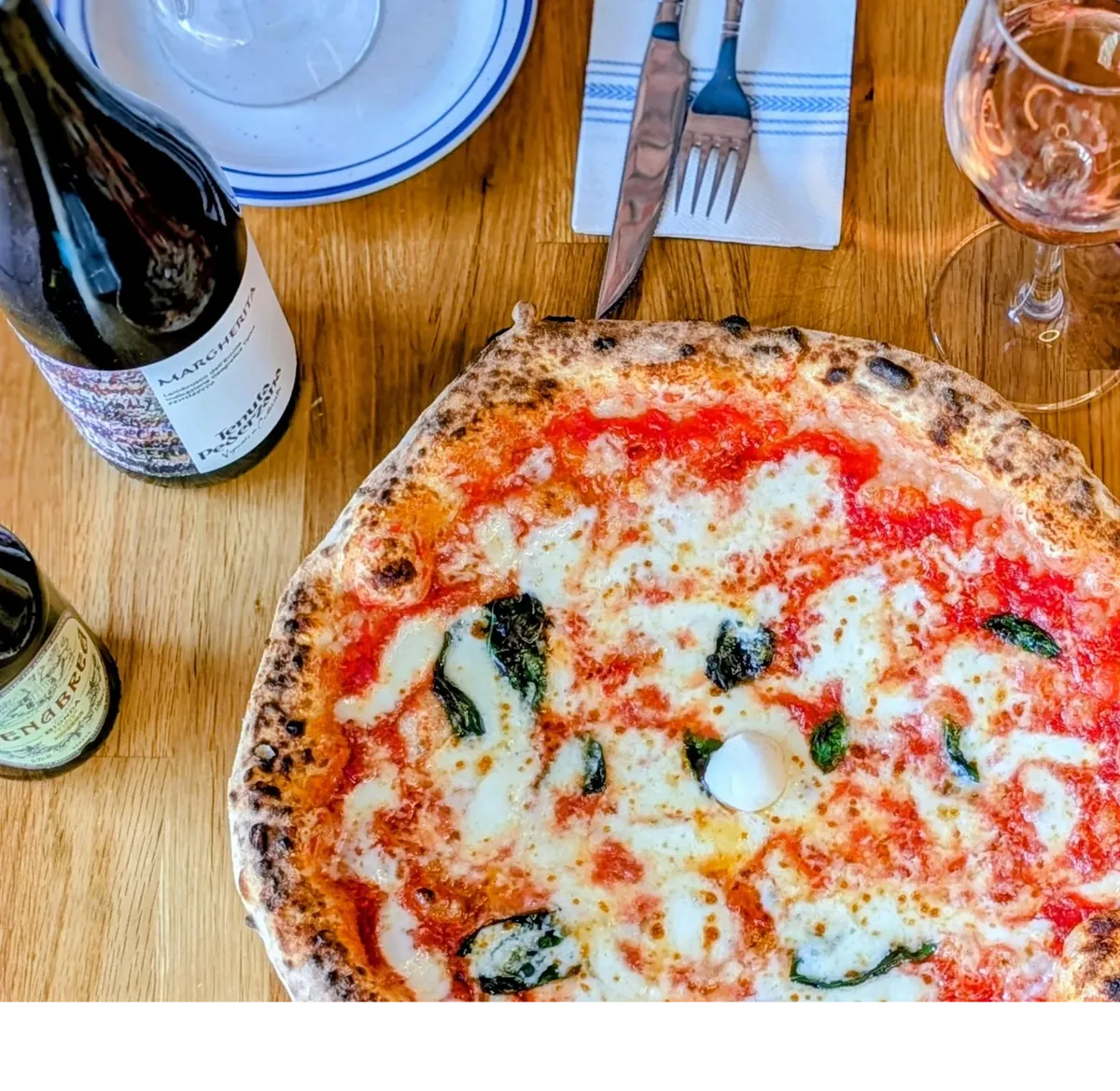 Margherita pizza, wine, and cutlery on a wooden table.