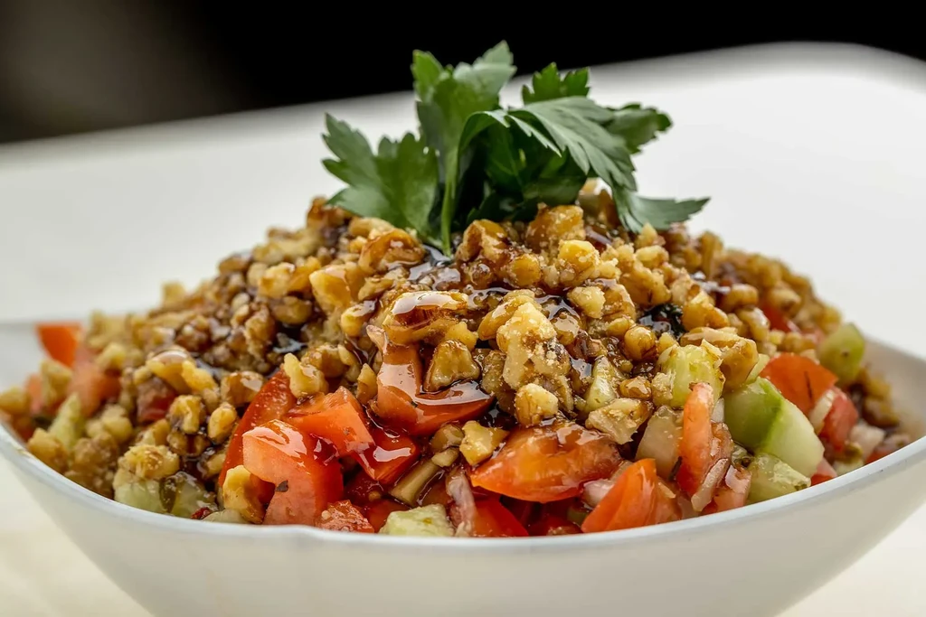 A white bowl of salad with chopped tomatoes, cucumbers, walnuts, and balsamic.