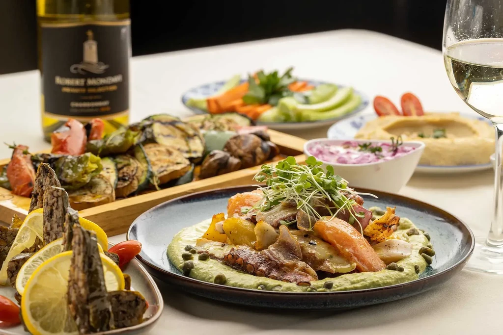 Table with roasted vegetables, seafood, dips, and white wine.