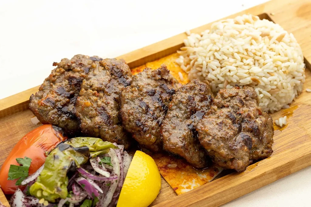 A wooden board with grilled meatballs, rice, salad, and lemon wedge.