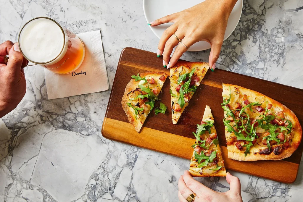 Hands holding a beer and flatbread pizza with arugula on a marble table.