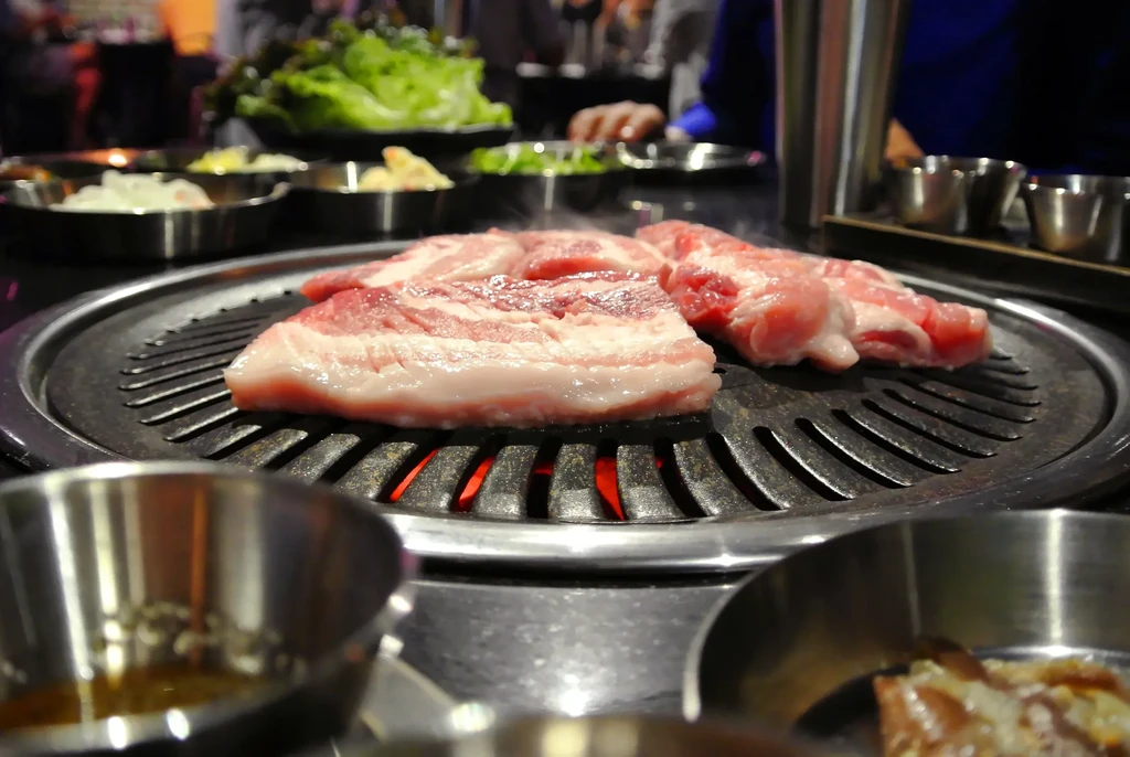 Pork belly strips cook on a Korean BBQ grill with red coals.