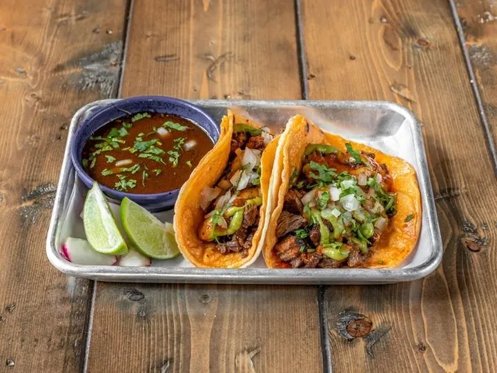 Two tacos, a bowl of soup, and lime slices on a metal tray.