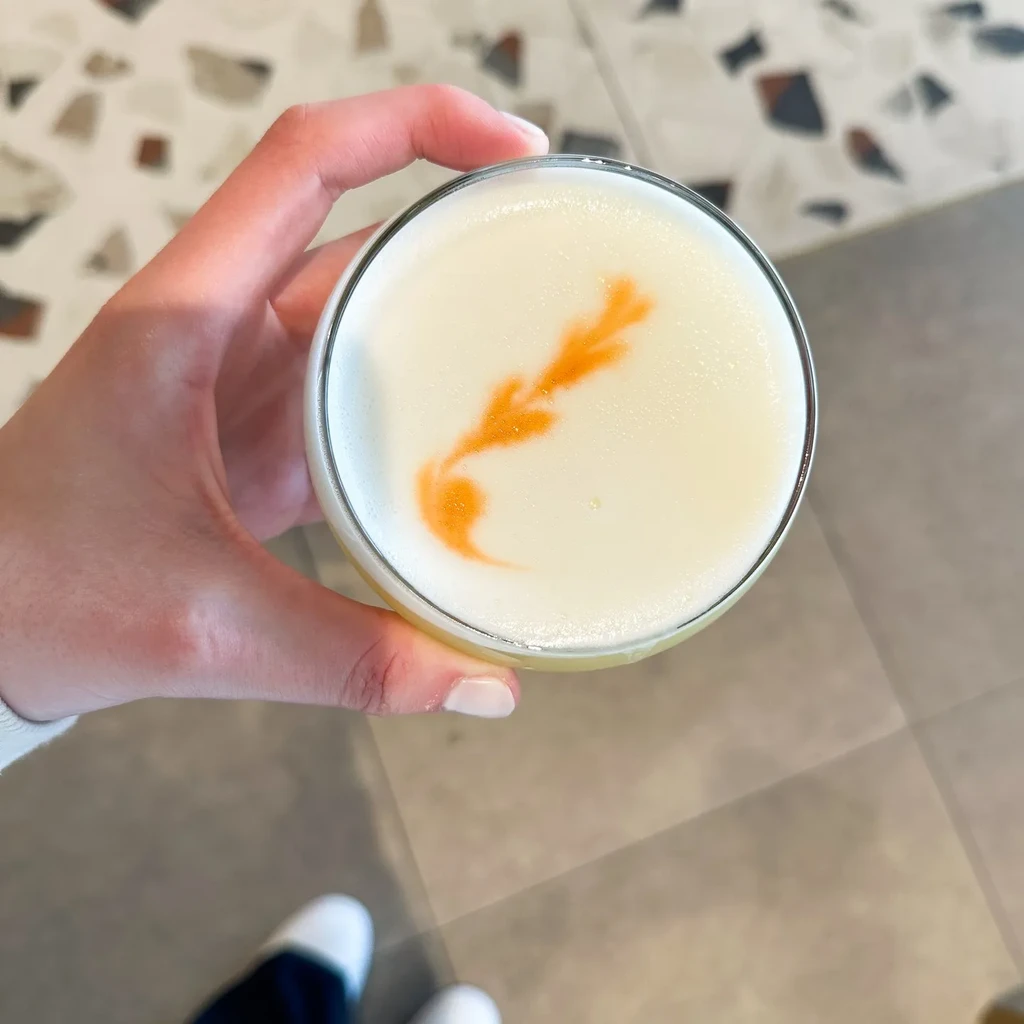 Hand holds cocktail with white foam and orange decorative drizzle.
