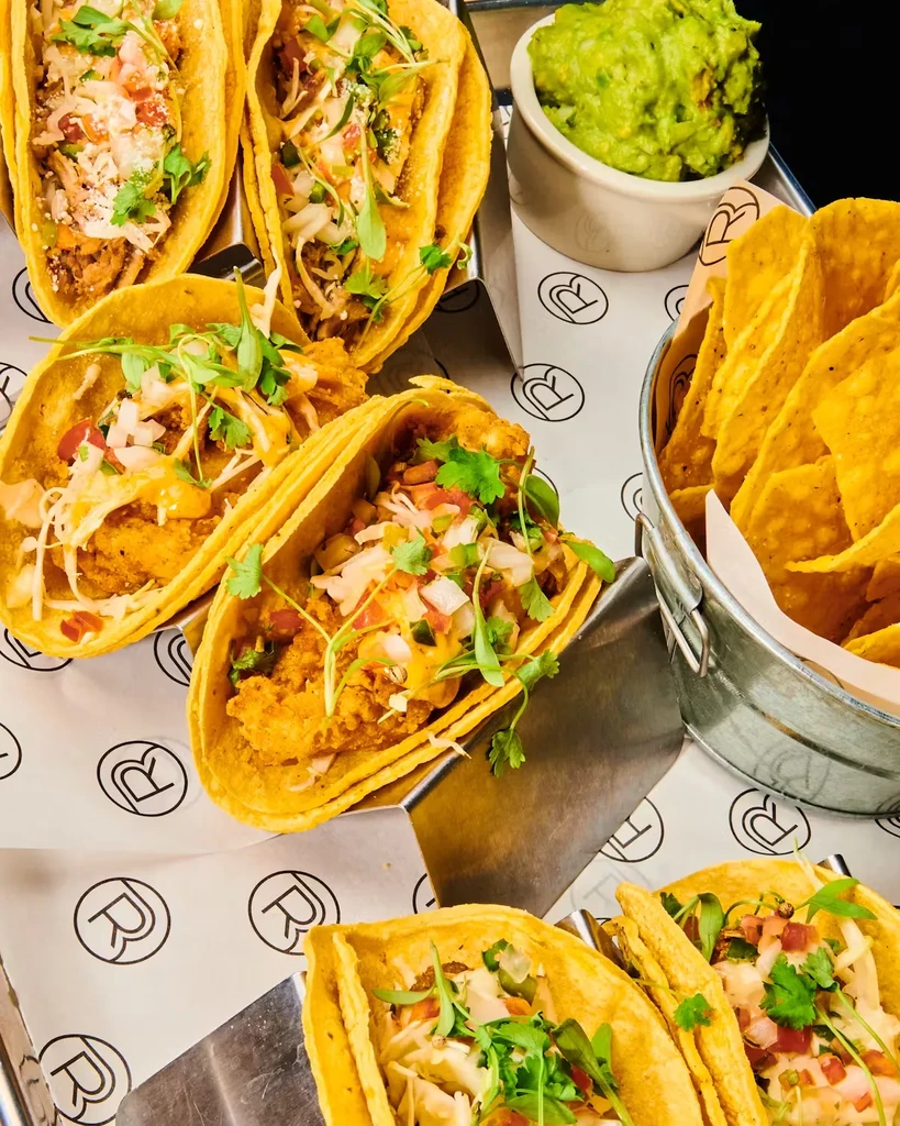 A variety of tacos, guacamole, and a bucket of tortilla chips.