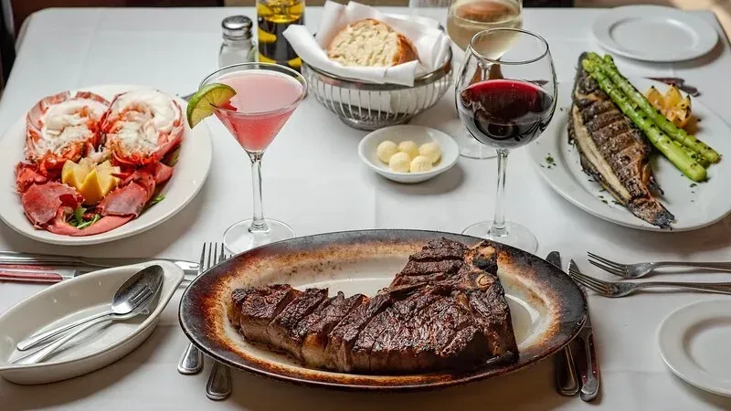 A large steak, lobster, fish, and drinks on a white tablecloth.