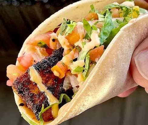 A hand holds a taco with dark meat, lettuce, pico de gallo, and sauce.