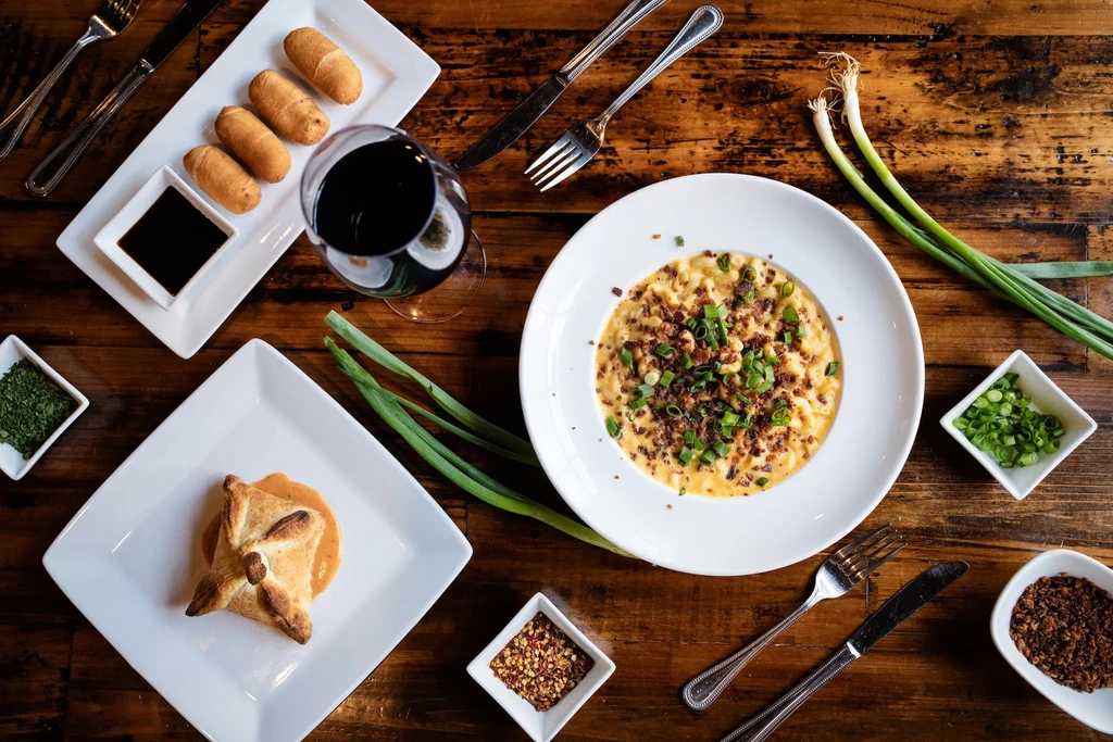 Flat lay of macaroni and cheese, pastries, wine, and garnishes on wood.