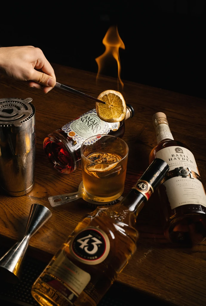 Hand torches orange peel over Amaro bottle, surrounded by spirits.