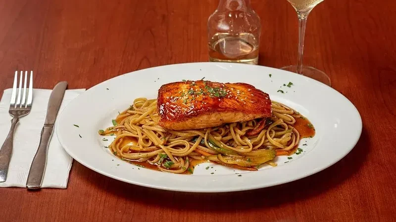 A plate of seared salmon over noodles with sauce on a wooden table.