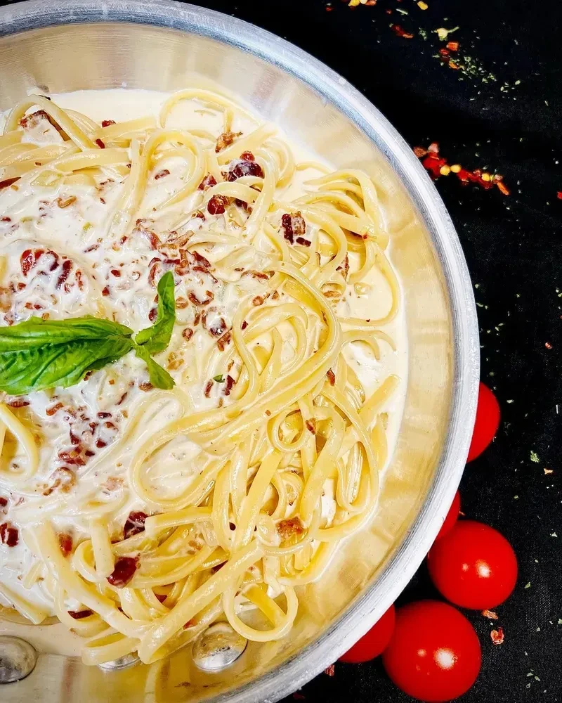 Creamy pasta with bacon bits and a basil leaf in a metal bowl.