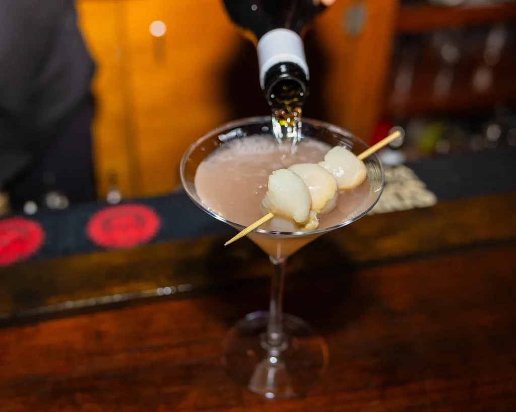 Bartender pouring liquid into martini glass with lychee fruit skewer.