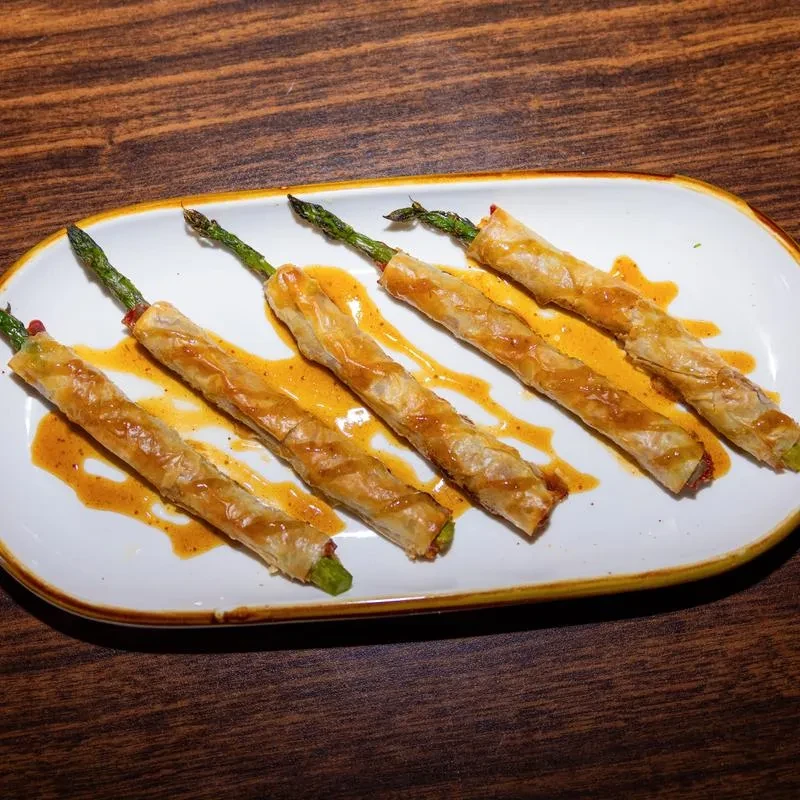 Six asparagus spears wrapped in pastry with sauce on a white plate.